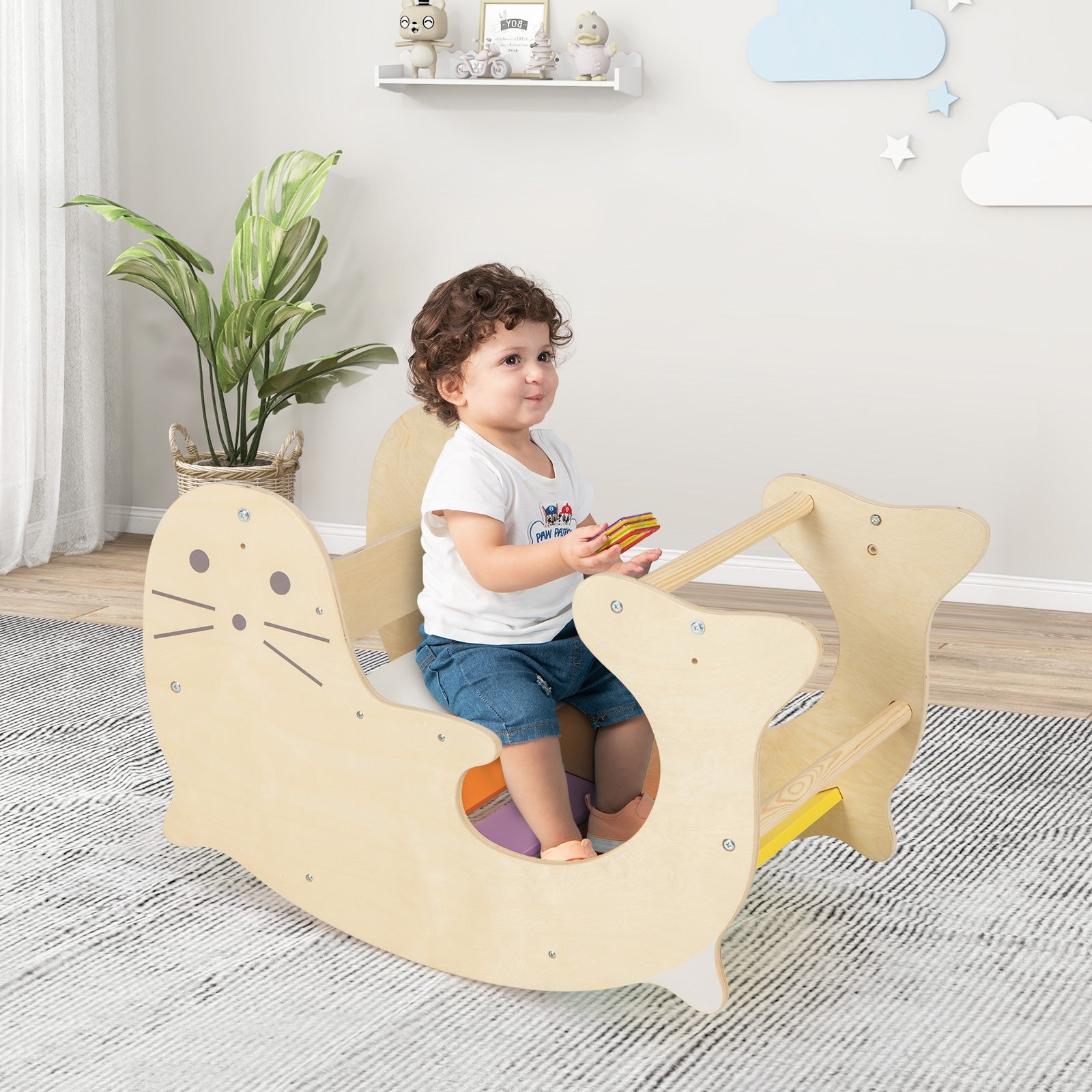 Child sitting on a wooden rocking toy in a room with a plant and shelves.